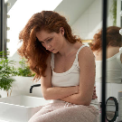 Kvinna med rött hår sitter i ett badrum och håller sig för magen